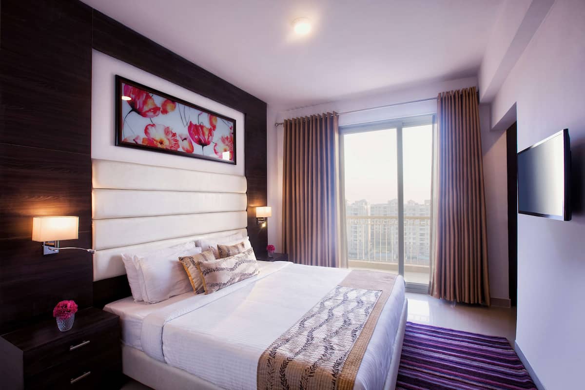 A modern hotel room with a large bed, a flat-screen TV, a window with curtains, and a city view.