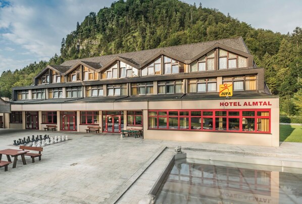 Experience the serene atmosphere of Hotel Almtal, surrounded by a forested hillside and a tranquil courtyard with a chessboard arrangement.