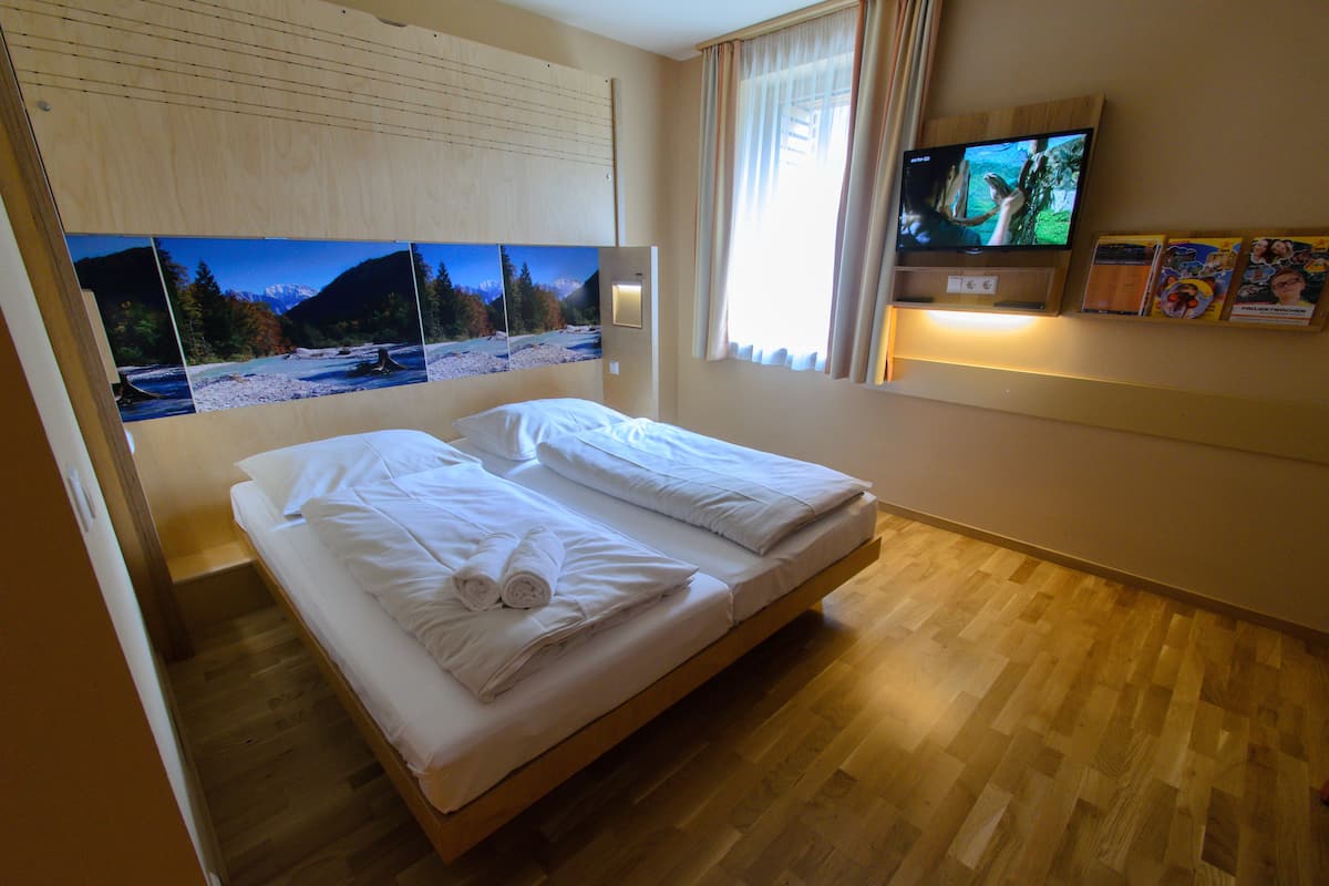 Habitación de hotel con cama, suelo de madera, televisor en la pared y ventana con cortinas.