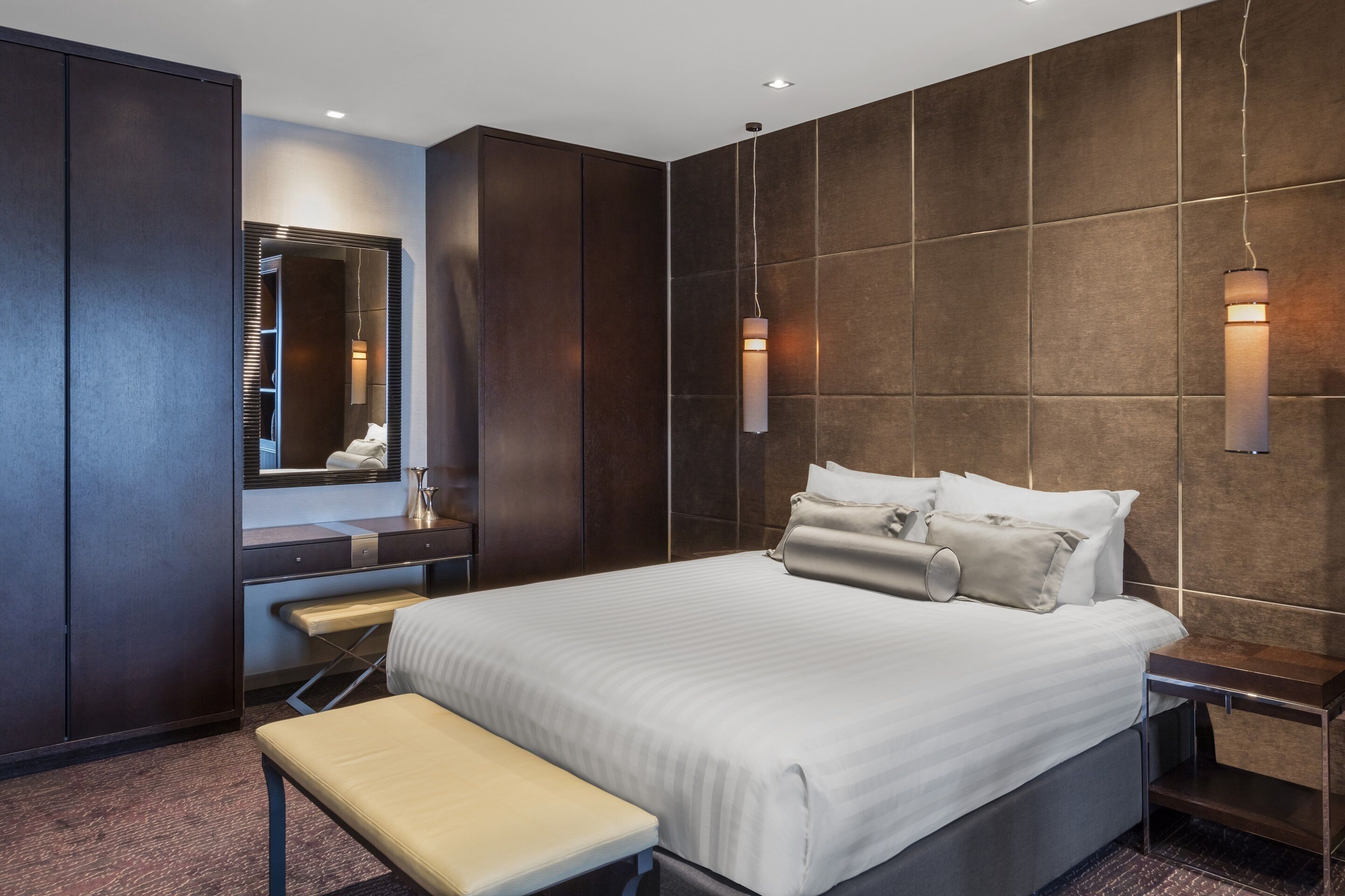 Une chambre d’hôtel moderne avec un grand lit, une banquette, une coiffeuse avec un miroir et deux lampes fixées au mur.
