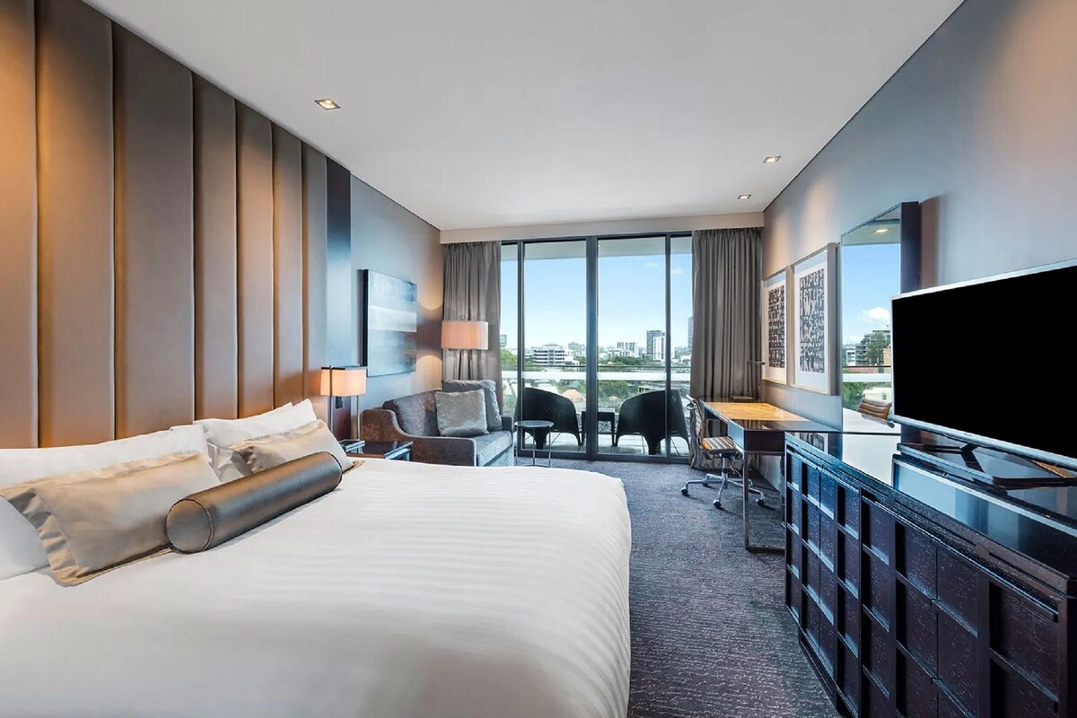 Una habitación de hotel moderna con una cama grande, un escritorio, una silla, un televisor y un balcón con vista a la ciudad.
