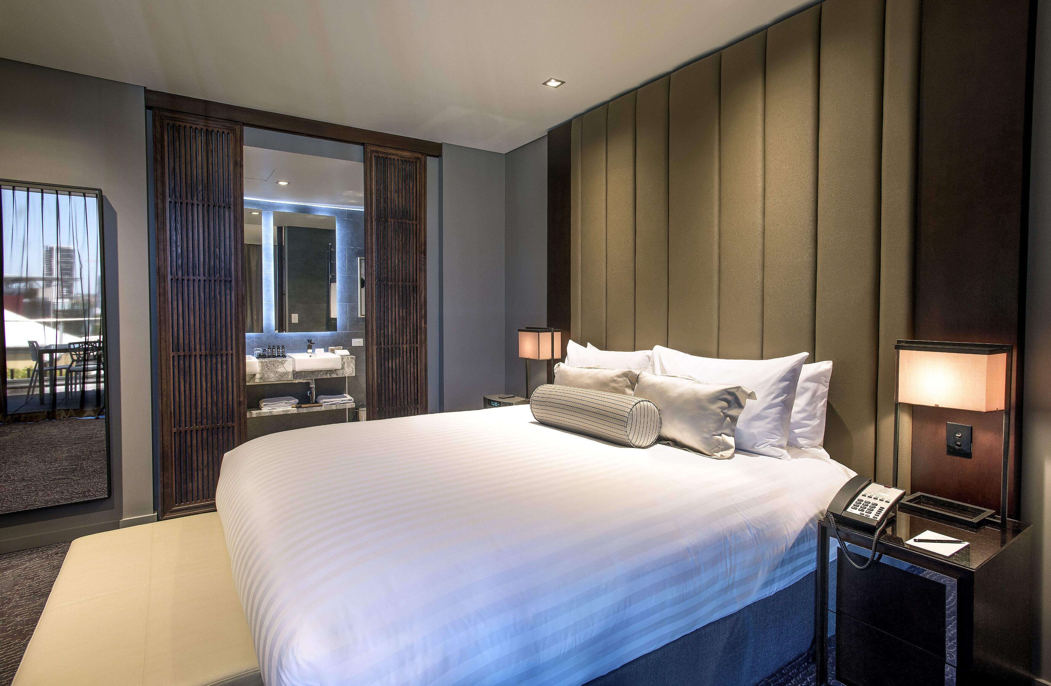 Habitación de hotel moderna con una cama grande, lámparas de noche, un teléfono y vistas de la ciudad a través de un ventanal amplio.