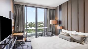 A modern hotel room with a large bed, a desk with a chair, a TV, and a balcony with a view of the city.