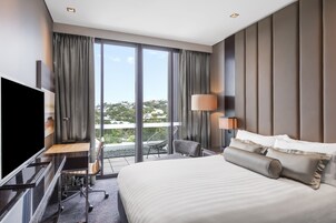 A modern hotel room with a large bed, a desk with a chair, a TV, and a balcony with a view of the city.
