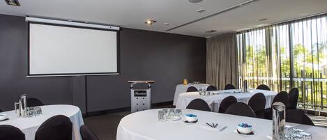 Uma sala de conferências com tela de projetor, mesas preparadas para uma reunião e vista para o exterior através de amplas janelas.
