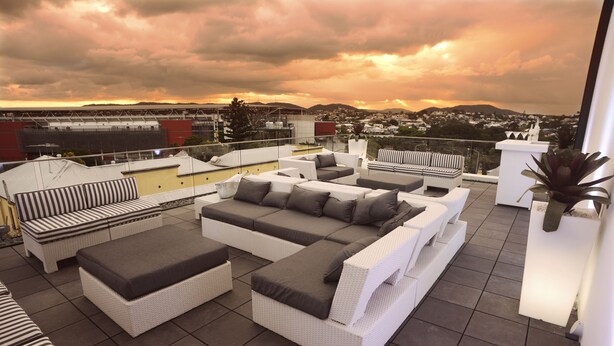 A rooftop terrace with white wicker furniture and a view of a cityscape at sunset. - The Beetson Hotel (Petrie Terrace)