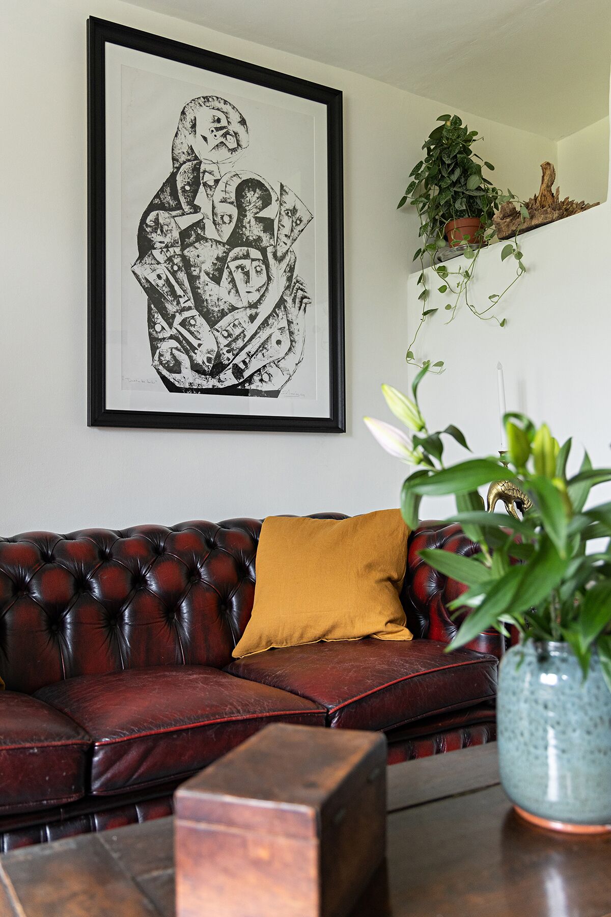 A dark red leather sofa with a mustard-colored pillow, a framed black and white artwork on the wall, and a potted plant on a wooden shelf.
