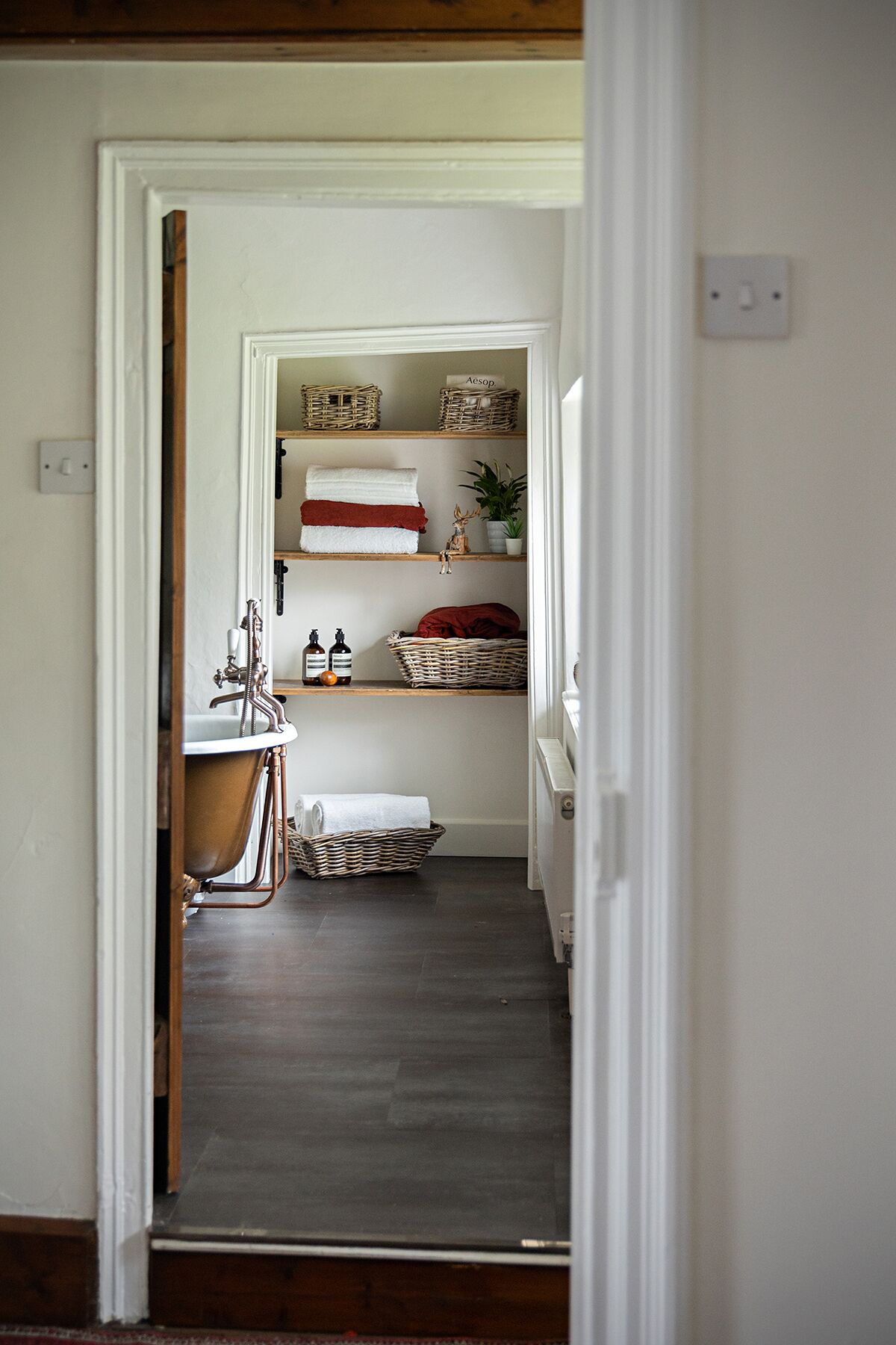 A narrow hallway with a vintage-style bathroom on the left, featuring a copper bathtub and a white sink. Shelves above hold baskets and a potted plant.