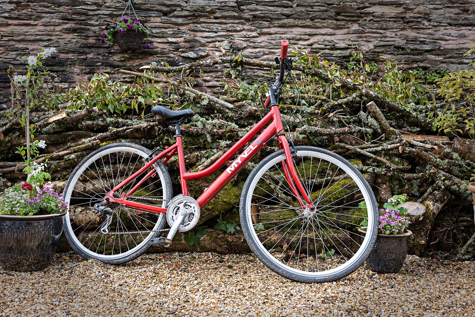 A red bicycle parked on gravel with a stone wall and potted plants in the background.