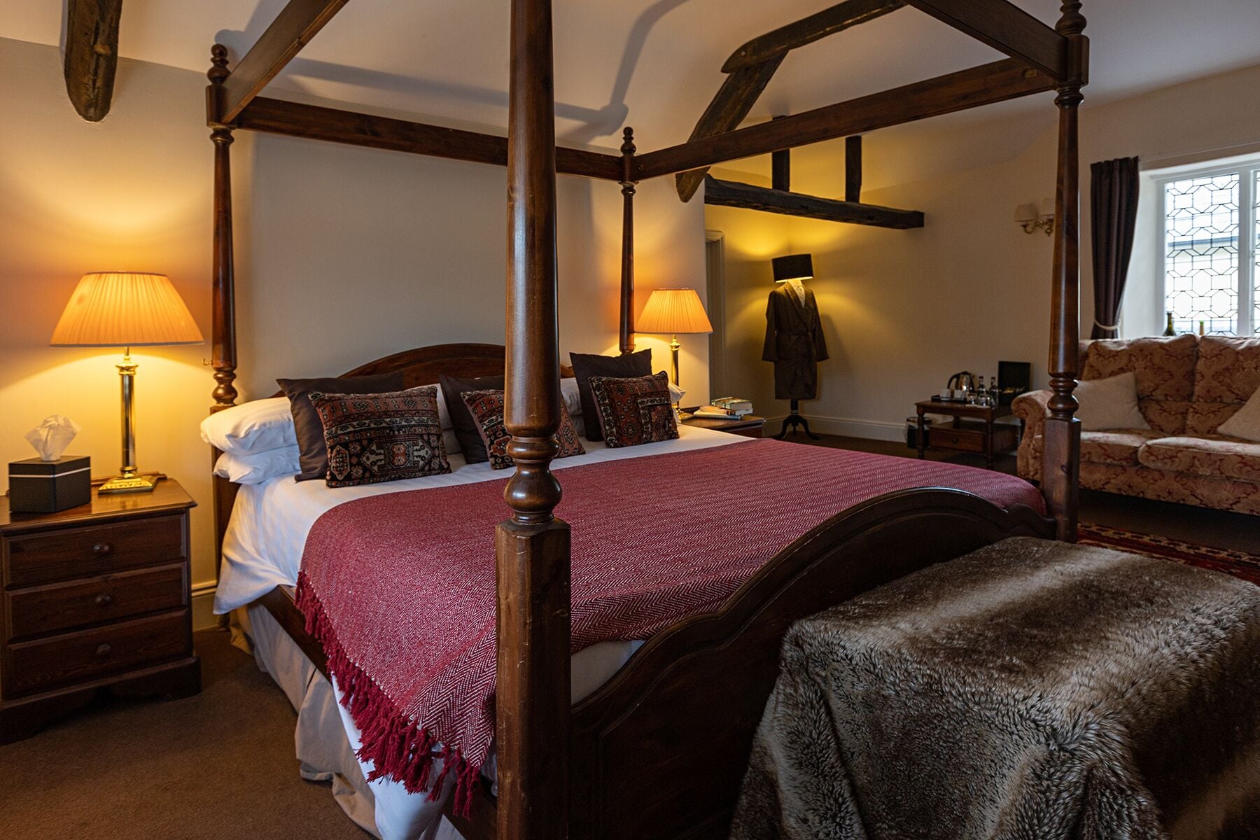 A four-poster bed with a red bedspread, a wooden nightstand with a lamp, a chair, and a robe hanging on the wall.