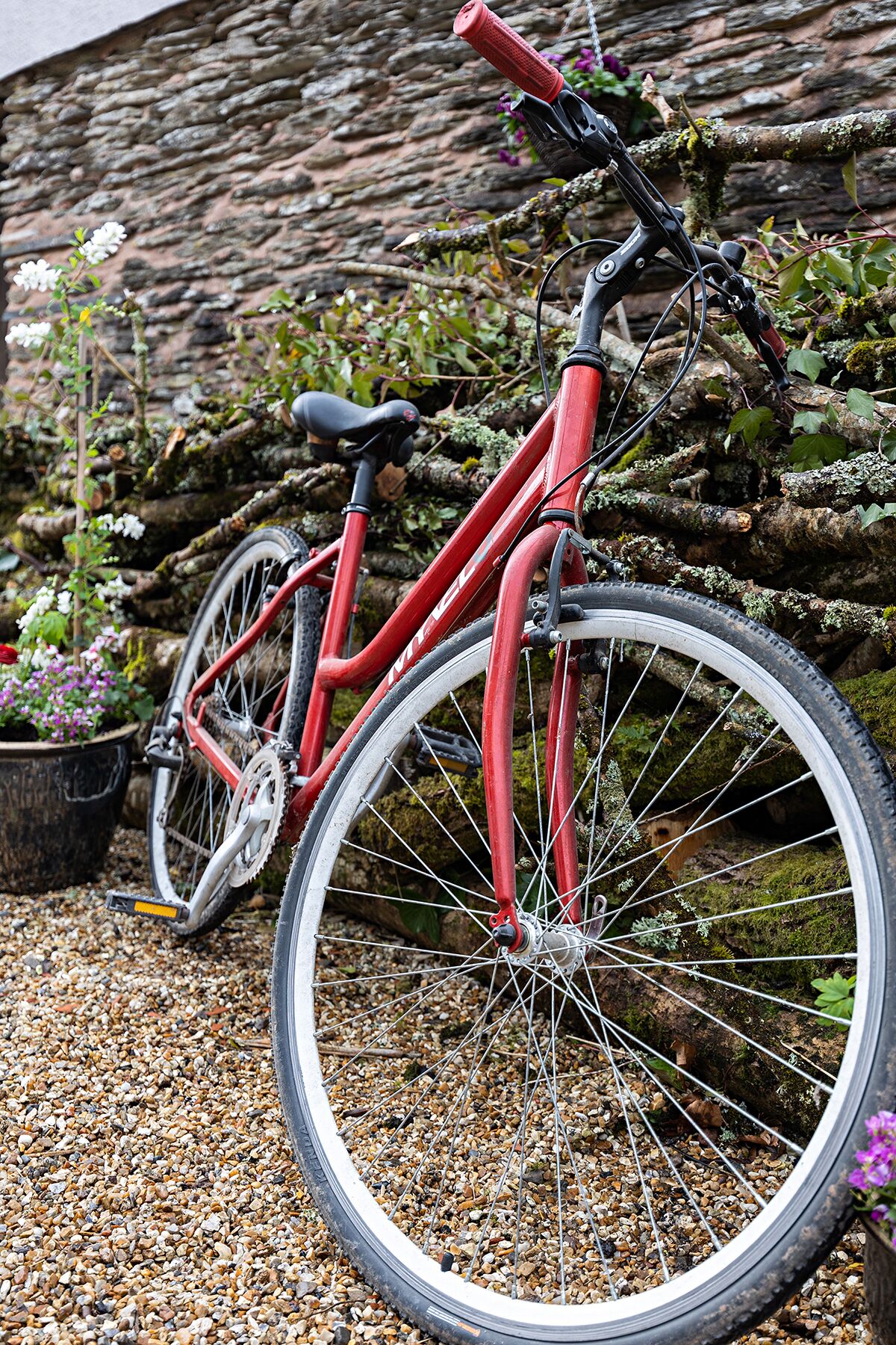 Un vélo rouge est garé sur un chemin de gravier, avec un mur de pierres et des plantes en arrière-plan.