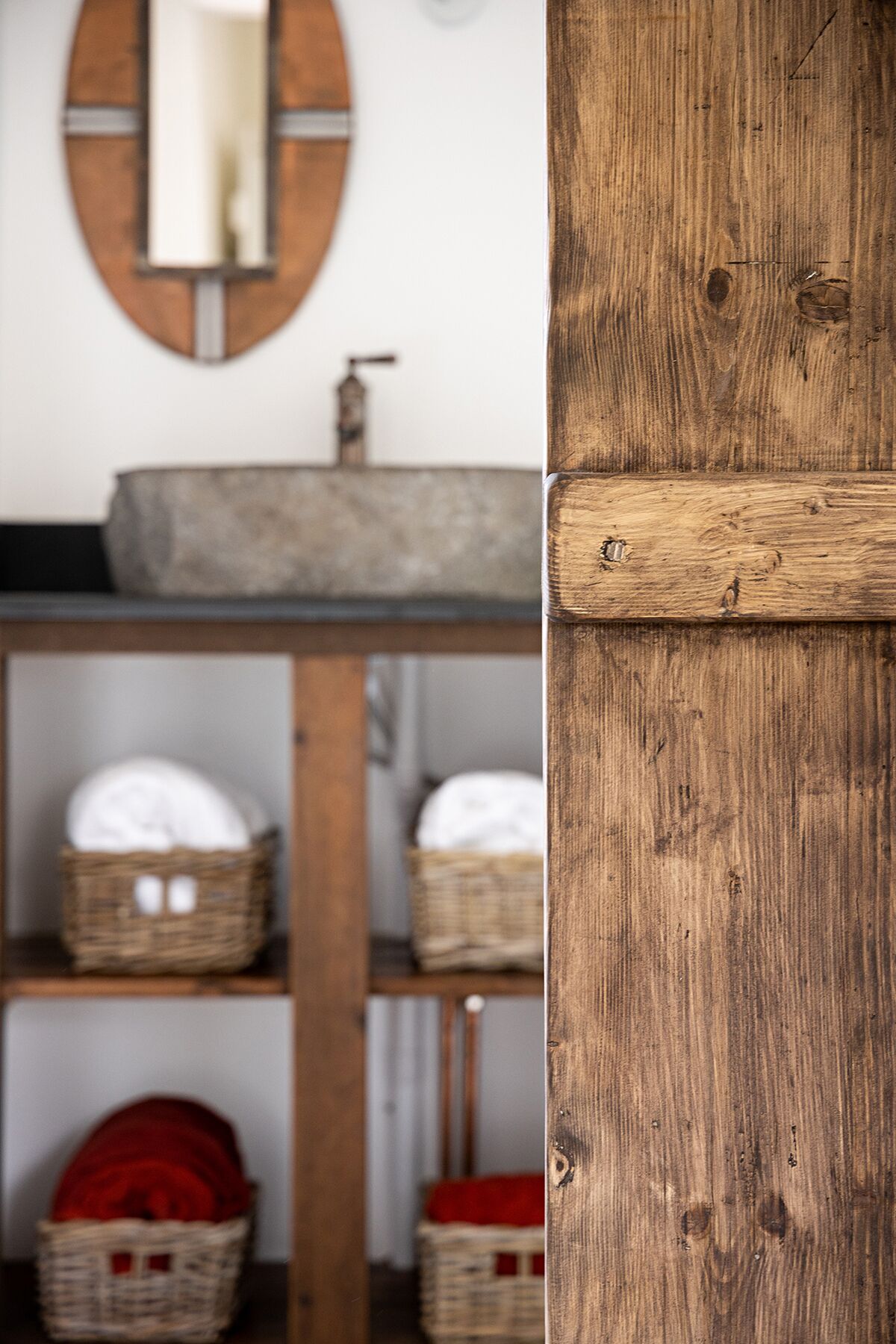 A rustic bathroom with a wooden vanity, a round mirror, and wicker baskets on shelves.
