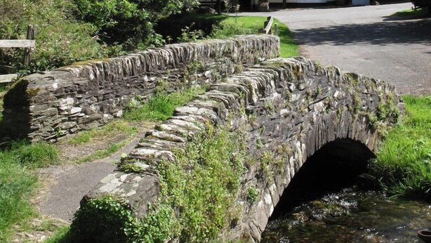A stone bridge over a stream in a rural setting with a traditional building in the background. - The Royal Oak Exmoor (Minehead)