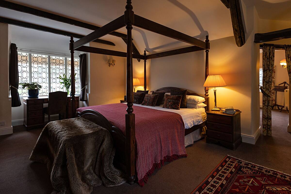 Ein Bett mit Baldachin, mit roter Bettdecke, ein Nachttisch mit Lampe und ein großes Fenster mit geometrischem Muster.
