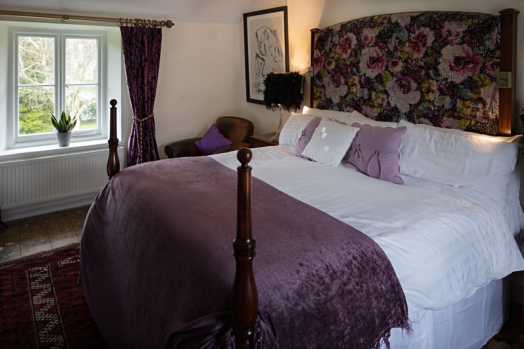 A bedroom with a large bed, a headboard with a floral tapestry, a window with curtains, and a potted plant.