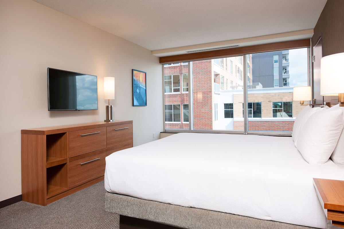 A hotel room with a large bed, a flat-screen TV, a wooden dresser, and a window with a view of buildings.