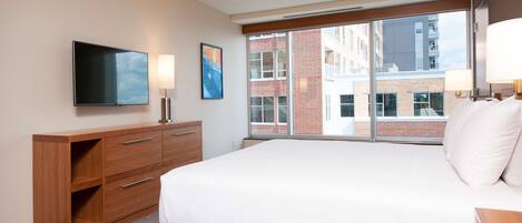 A hotel room with a large bed, a flat-screen TV, a wooden dresser, and a window with a view of buildings.