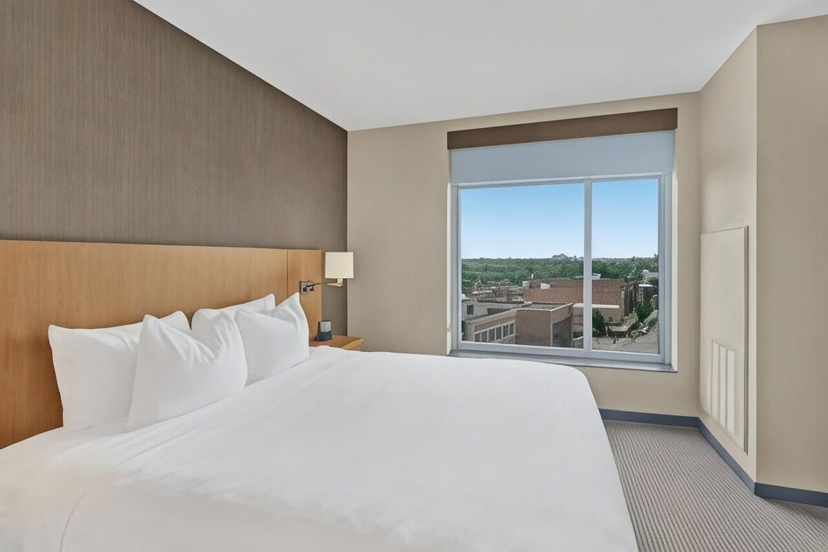 A hotel room with a large bed, a nightstand, a lamp, and a large window with a city view.