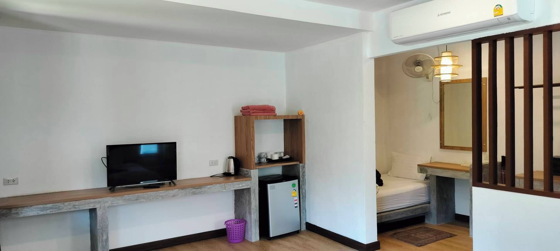 Ein kleines Zimmer mit Fernseher auf einer Holzablage, einem kleinen Kühlschrank, einem Holfachschrank mit Küchenutensilien, einem Bett mit Holz Kopfteil und einem Spiegel über dem Bett.