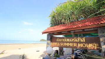 A beachside summer house with a sign in Thai, a palm tree, and beach chairs.