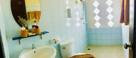 Un baño con lavamanos, inodoro y ducha. Hay un perchero con toallas y un espejo decorativo.