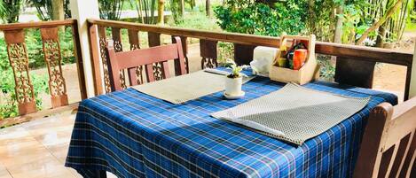 A table set with a blue checkered tablecloth, two placemats, a small potted plant, and a container with various items on a balcony with a view of lush greenery.
