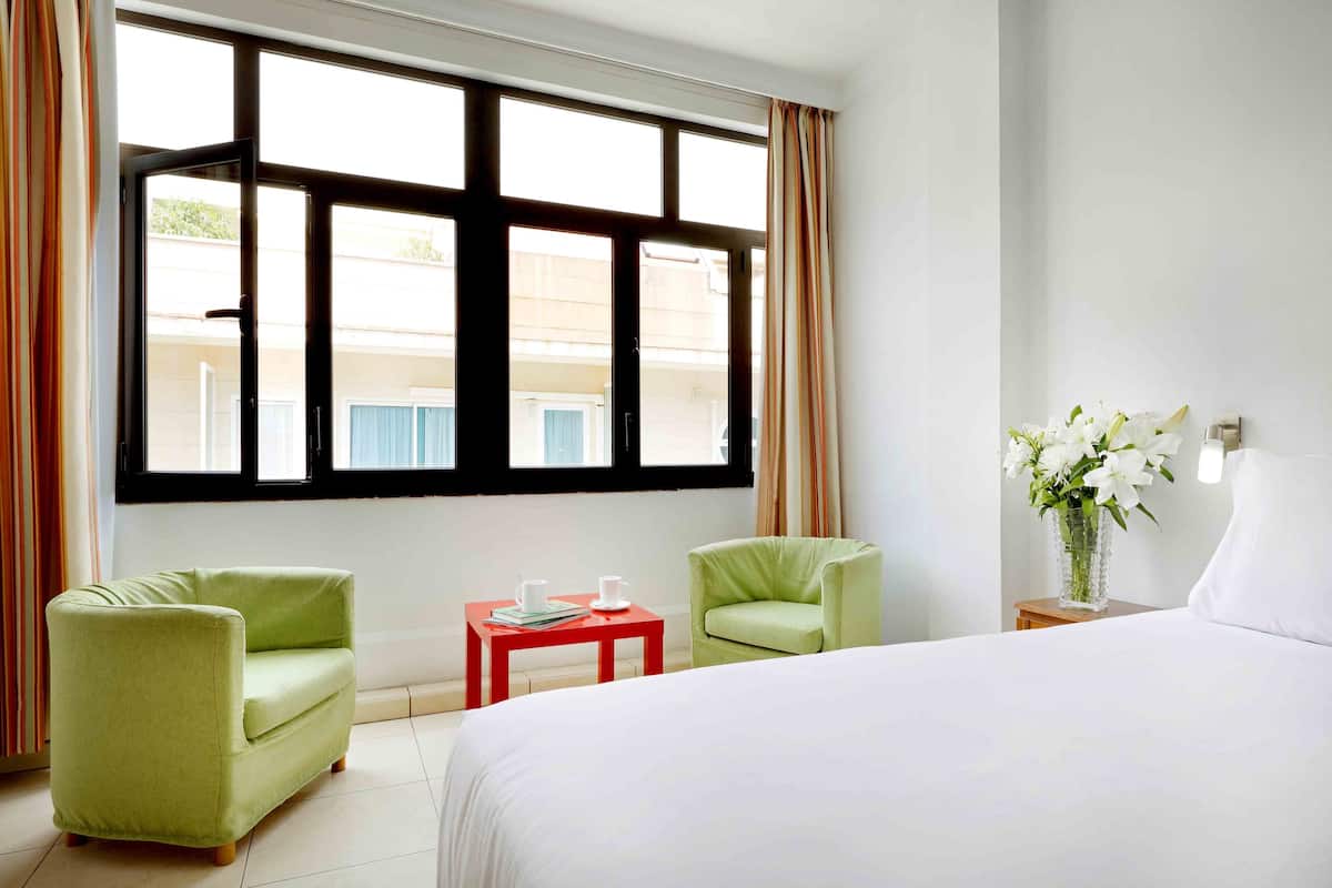 Een hotelkamer met een groot bed, twee groene fauteuils, een kleine rode tafel met een witte bloevas, en een raam met gordijnen.
