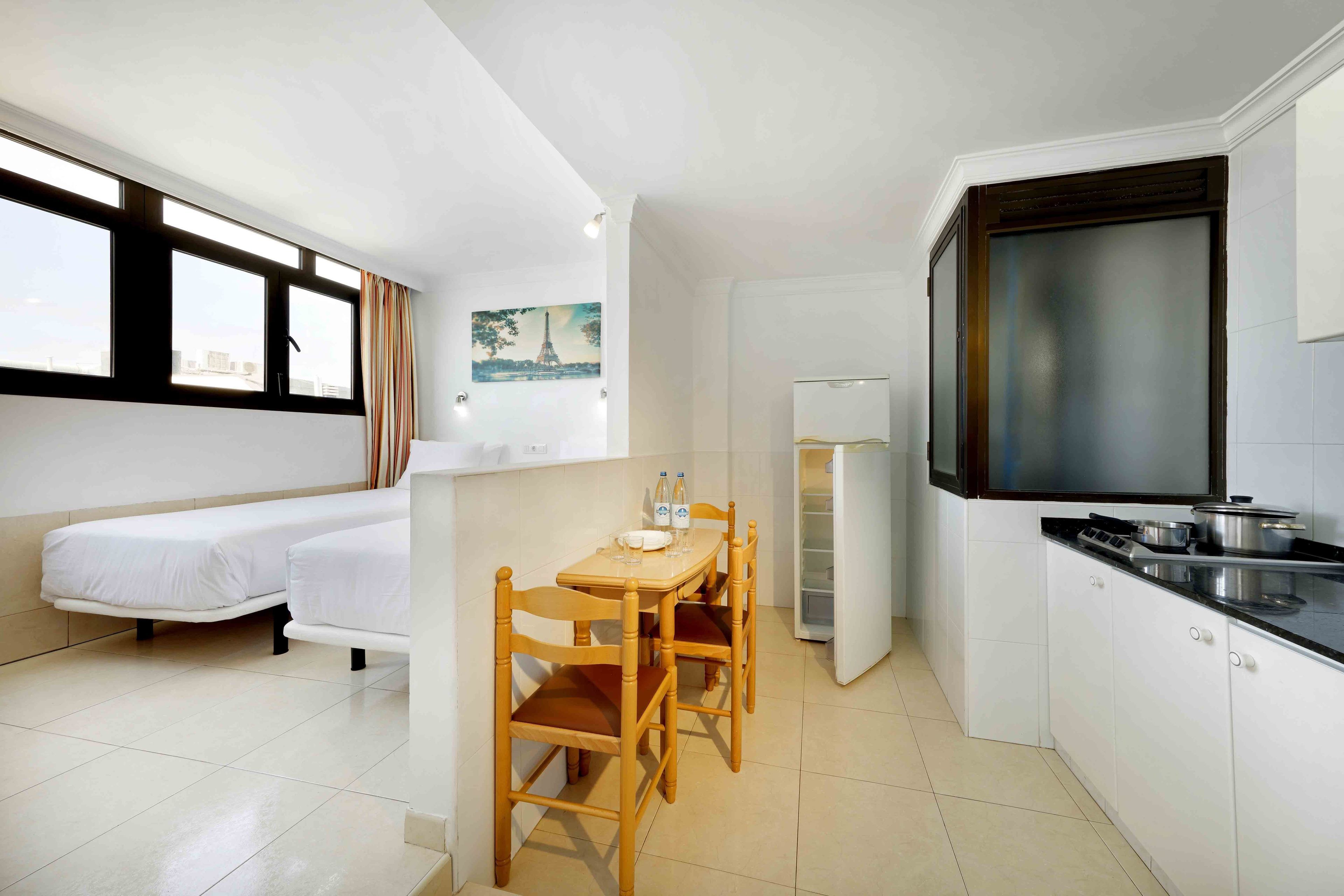 A compact hotel room with a kitchenette, a dining area with a table and chairs, a bed, and a window with curtains.