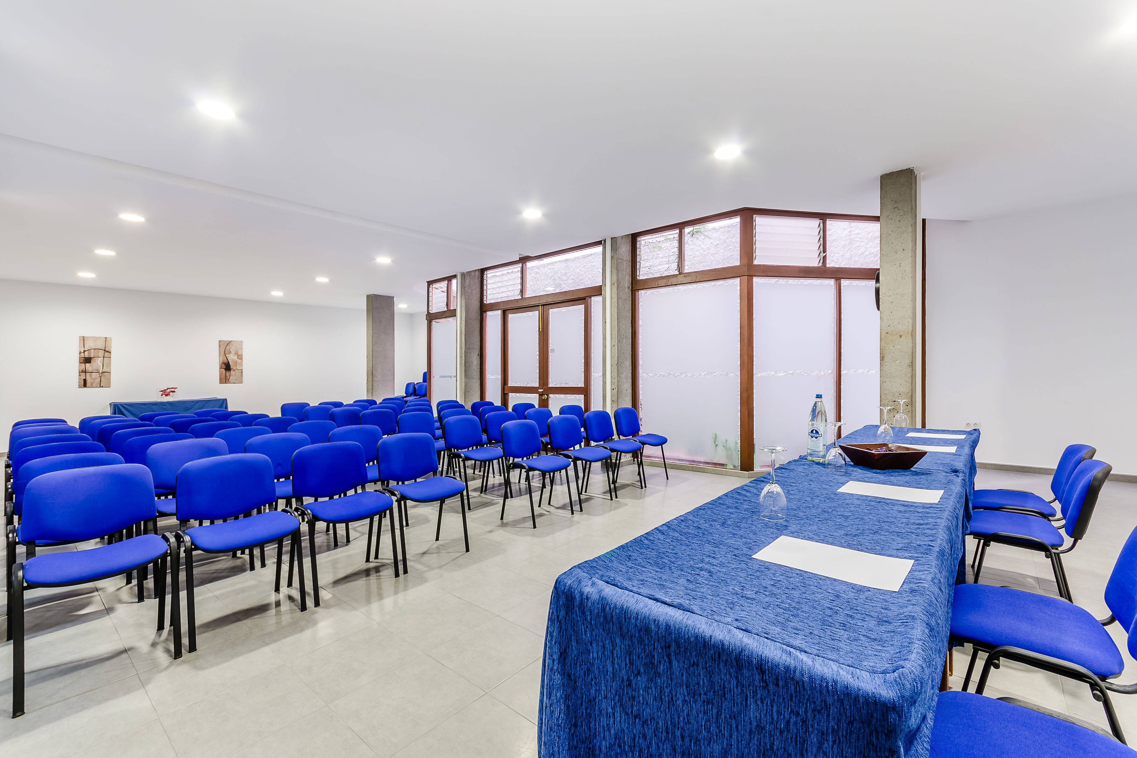 A conference room with blue chairs and a long table, white walls, and large windows.