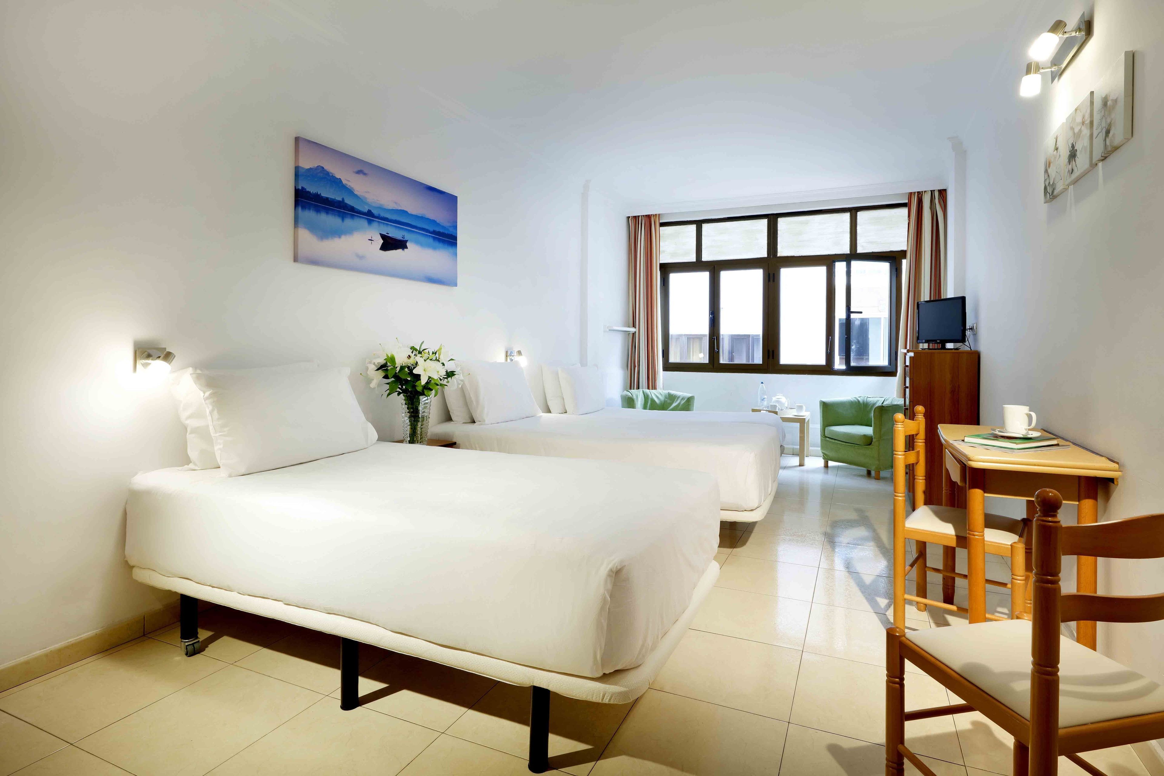 A hotel room with two beds, a small table with a chair, a TV, and a window with curtains.