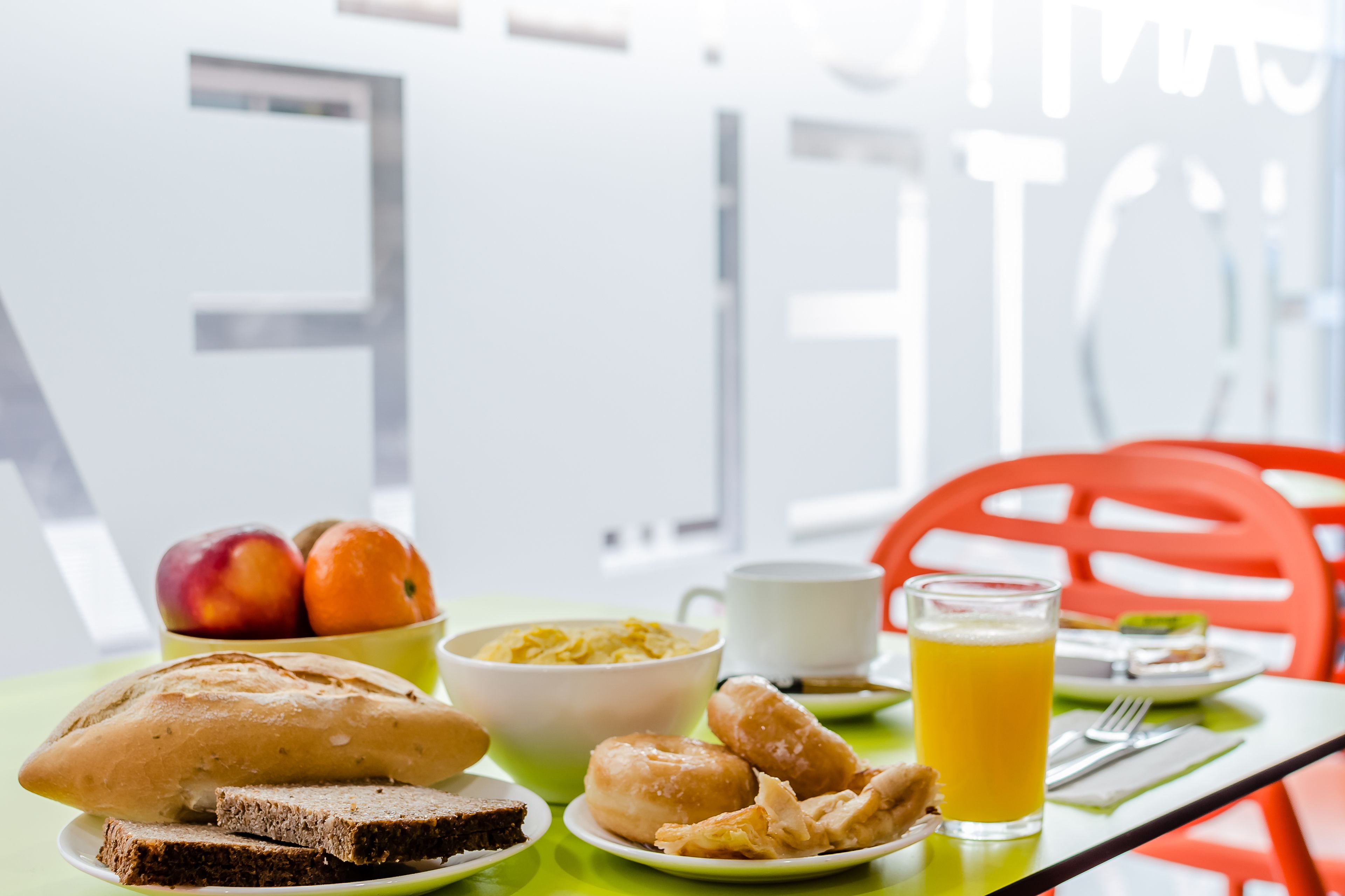 Mesa de café da manhã com pão, doces, frutas, uma tigela de cereal, uma xícara e um copo de suco de laranja.
