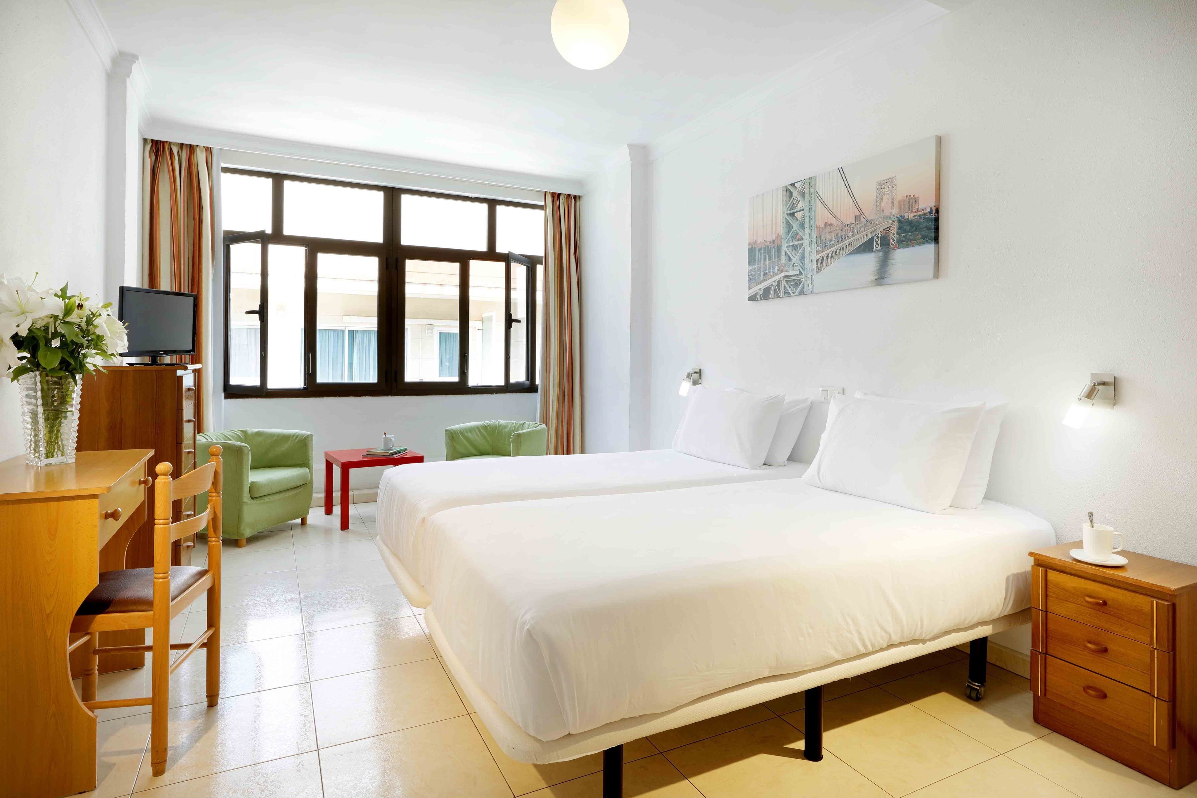 Habitación de hotel con una cama grande, un escritorio con silla, televisor, una mesita y ventana con cortinas.