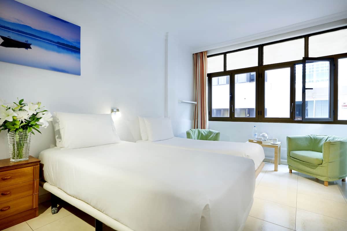 Hotelkamer met een groot bed, een nachtkastje, een groene fauteuil, een tafeltje met een waterfles en een beker, en een raam met gordijnen.