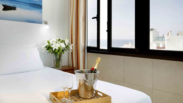Hotelkamer met een bed, een raam met uitzicht op de stad, een minibar met een fles champagne en een dienblad met glazen.