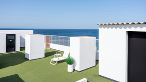 Een terras op het dak met uitzicht op zee, witte muren en een klein zitgedeelte met een plant.