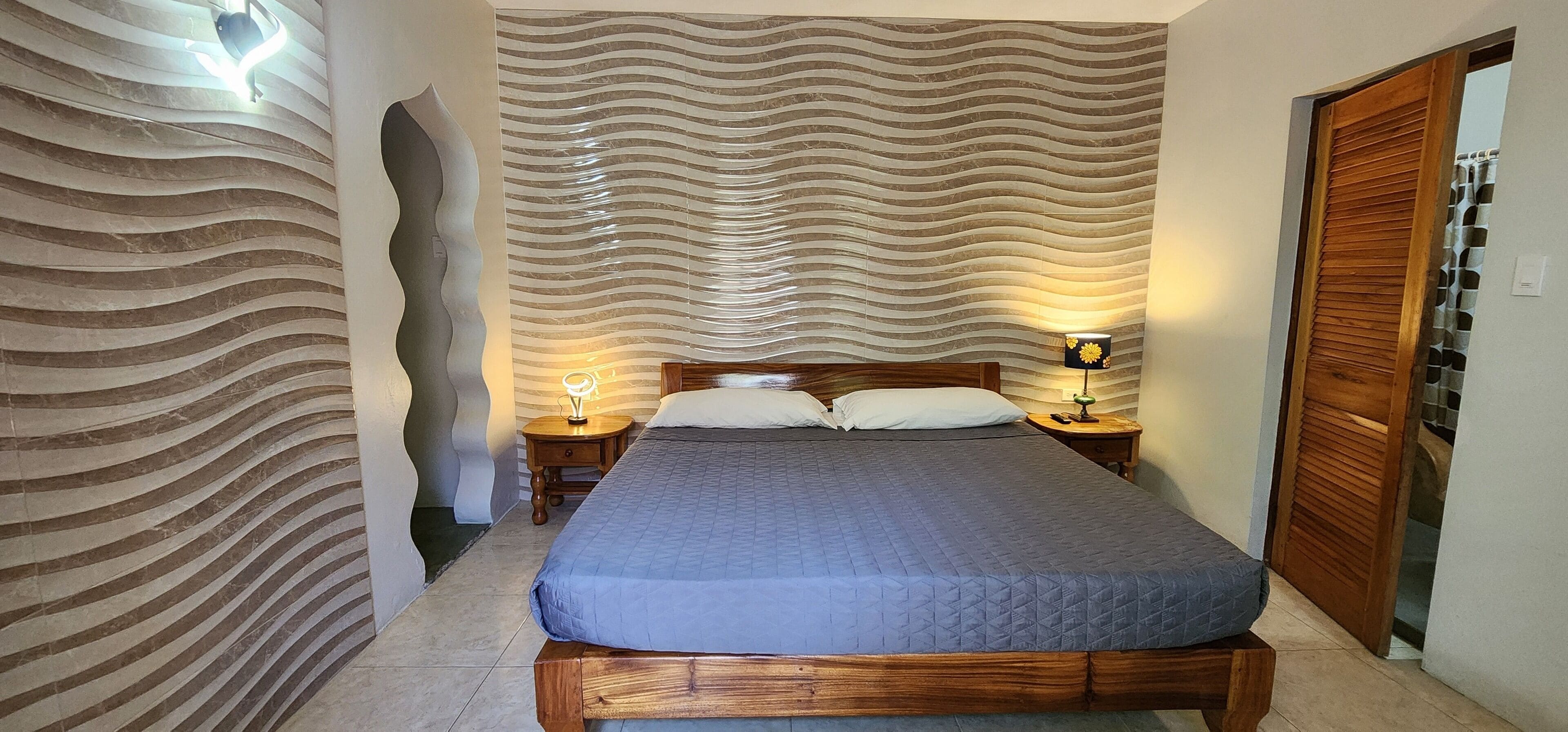 A bedroom with a wooden bed, two bedside tables with lamps, a patterned wall, and a wooden door.