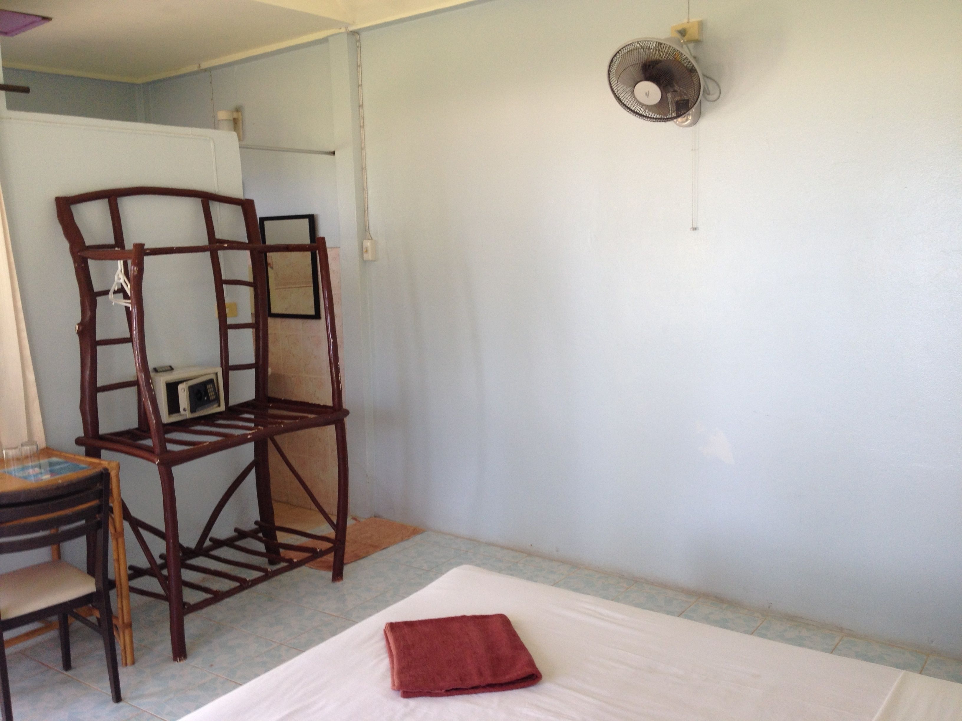 A room with a bed, a wooden chair, a small table, and a wall-mounted fan.