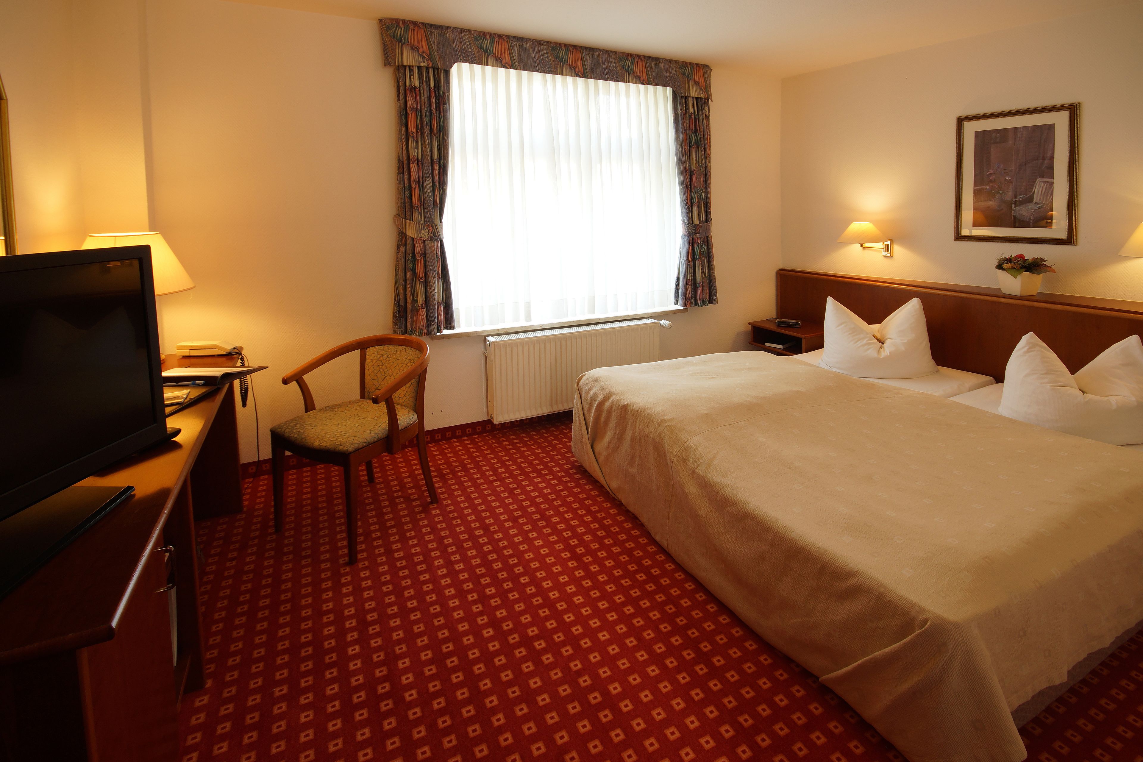 Une chambre d’hôtel avec un grand lit, un bureau avec une télévision, une chaise, une fenêtre avec des rideaux, une table de chevet et une lampe fixée au mur.