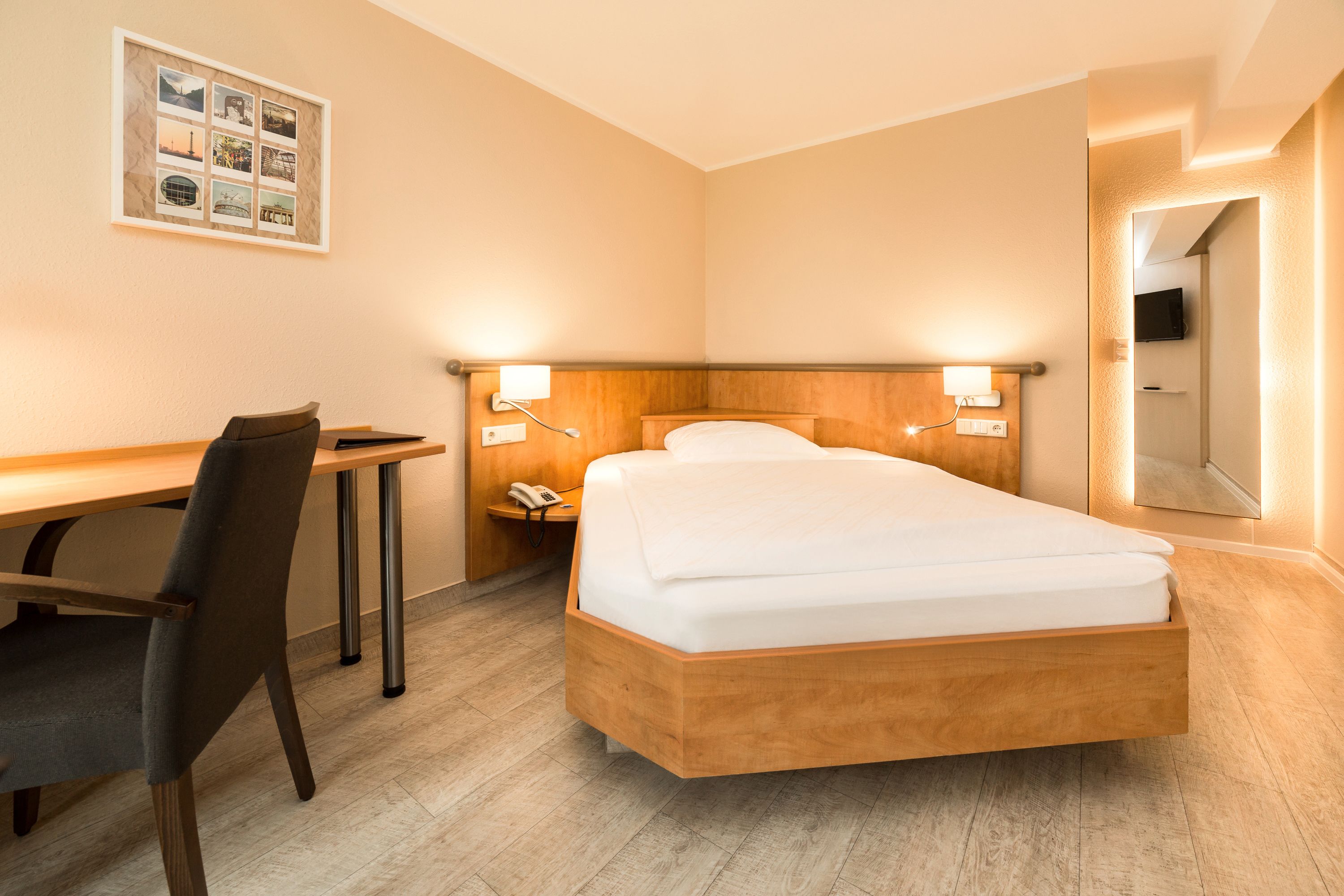 Hotellihuone, jossa on puinen sänky, yöpöytä lampun kanssa, työpöytä tuolilla ja seinällä kehystetty valokuva.