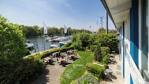 Un patio a la orilla del río con mesas y sillas, con vista a un puerto deportivo con barcos.