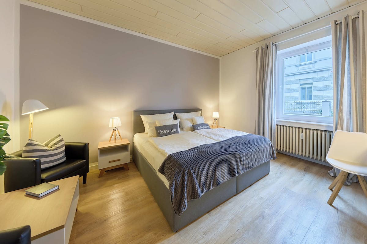 A modern bedroom with a large bed, bedside tables with lamps, a chair, and a view of the city through a window.