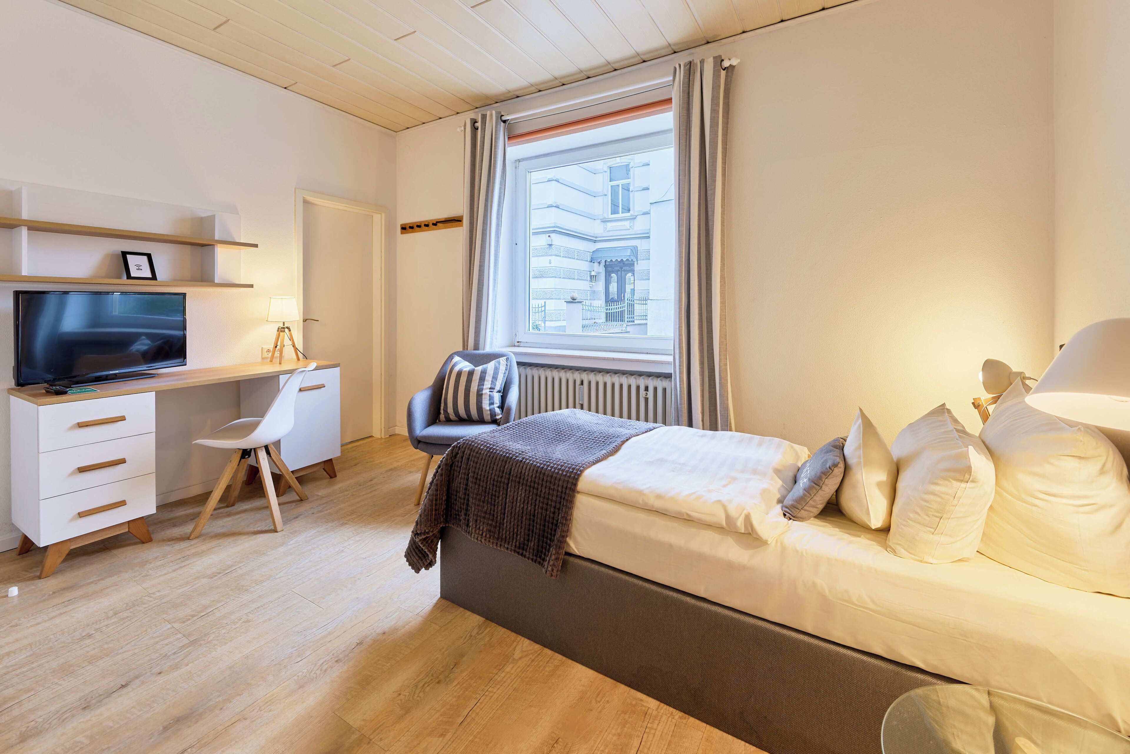 A modern bedroom with a bed, a desk, a chair, a TV, and a window with curtains.