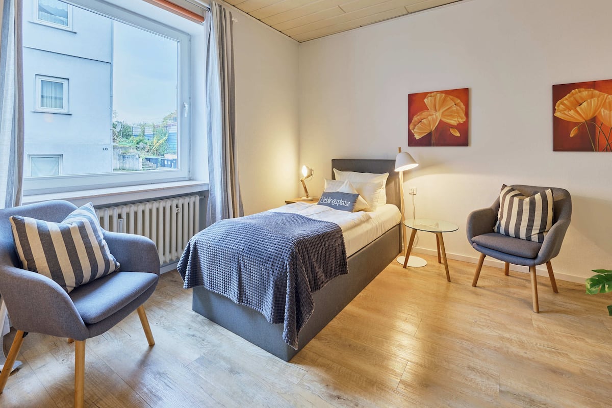 A modern bedroom with a bed, two chairs, a small table, and artwork on the wall.