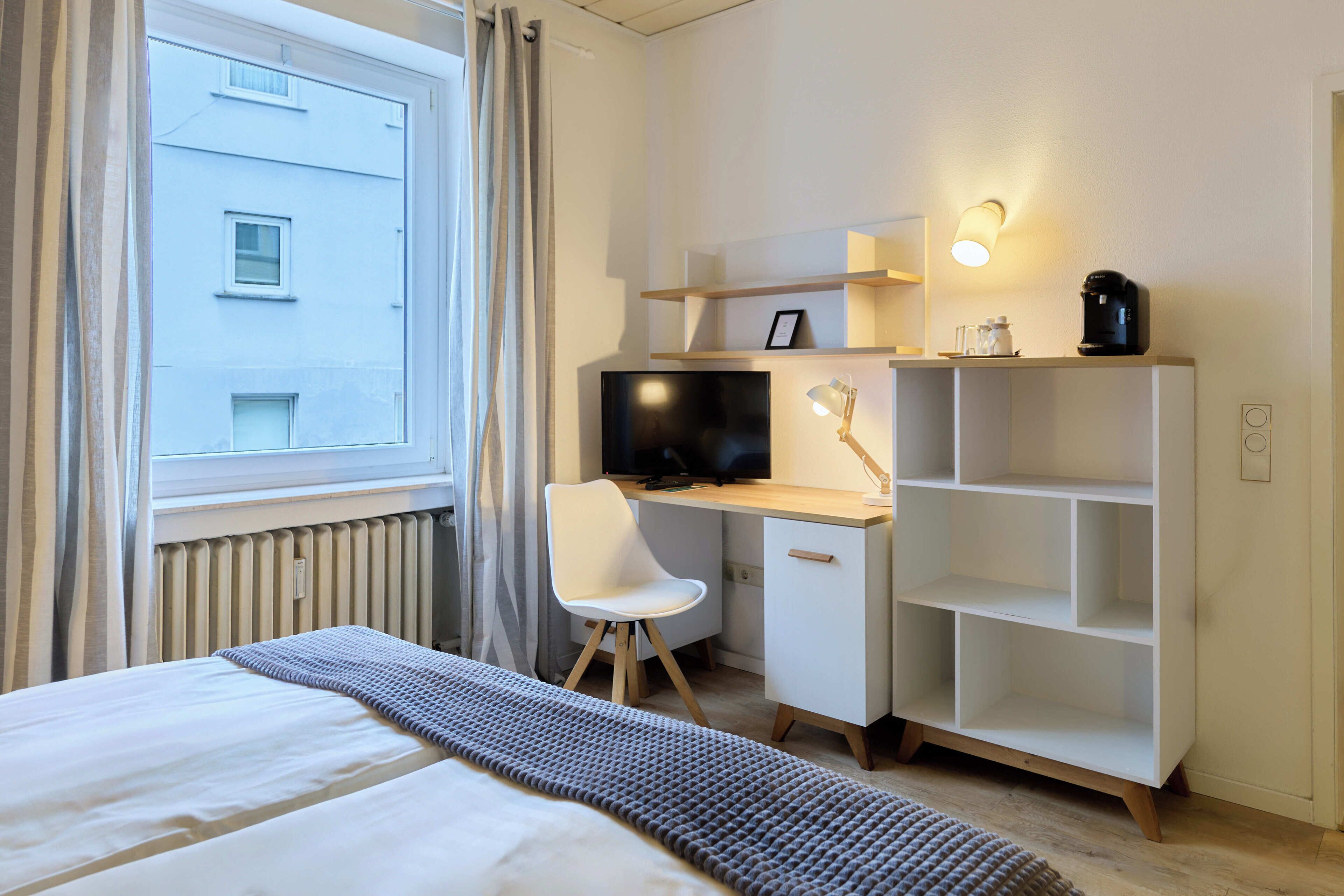 A modern hotel room with a bed, a desk with a computer, a chair, a TV, and a shelf with a coffee maker.