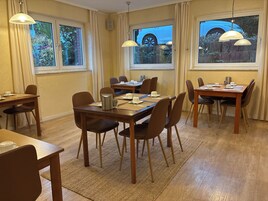 Un comedor con varias mesas y sillas, ventanas amplias con cortinas y vistas al exterior con coches y vegetación.