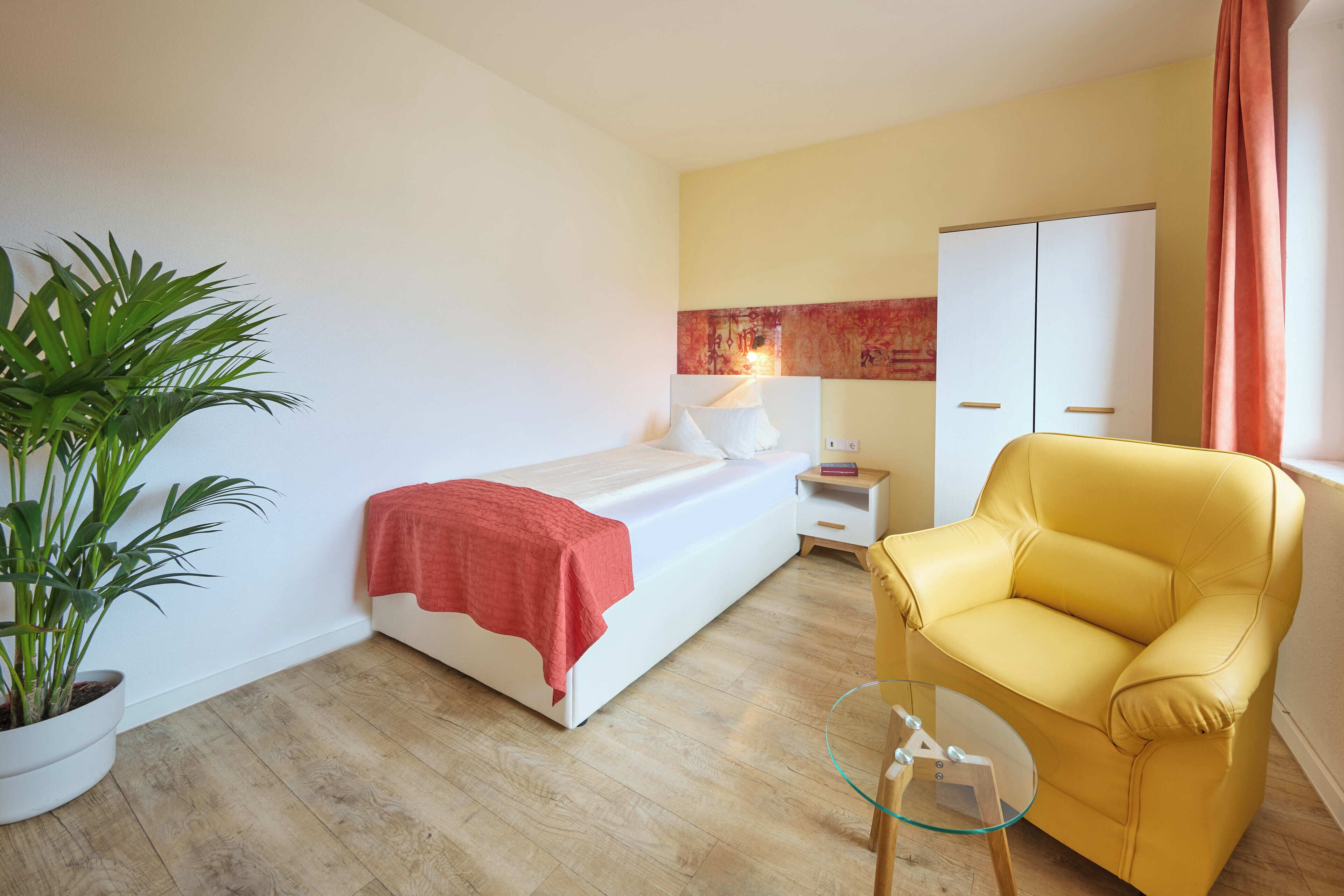 A hotel room with a bed, a nightstand, a chair, a small table, a wardrobe, and a potted plant.