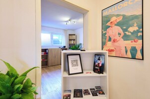 Zona de recepción con un código QR enmarcado, un cartel de Mallorca y una estantería blanca con libros.