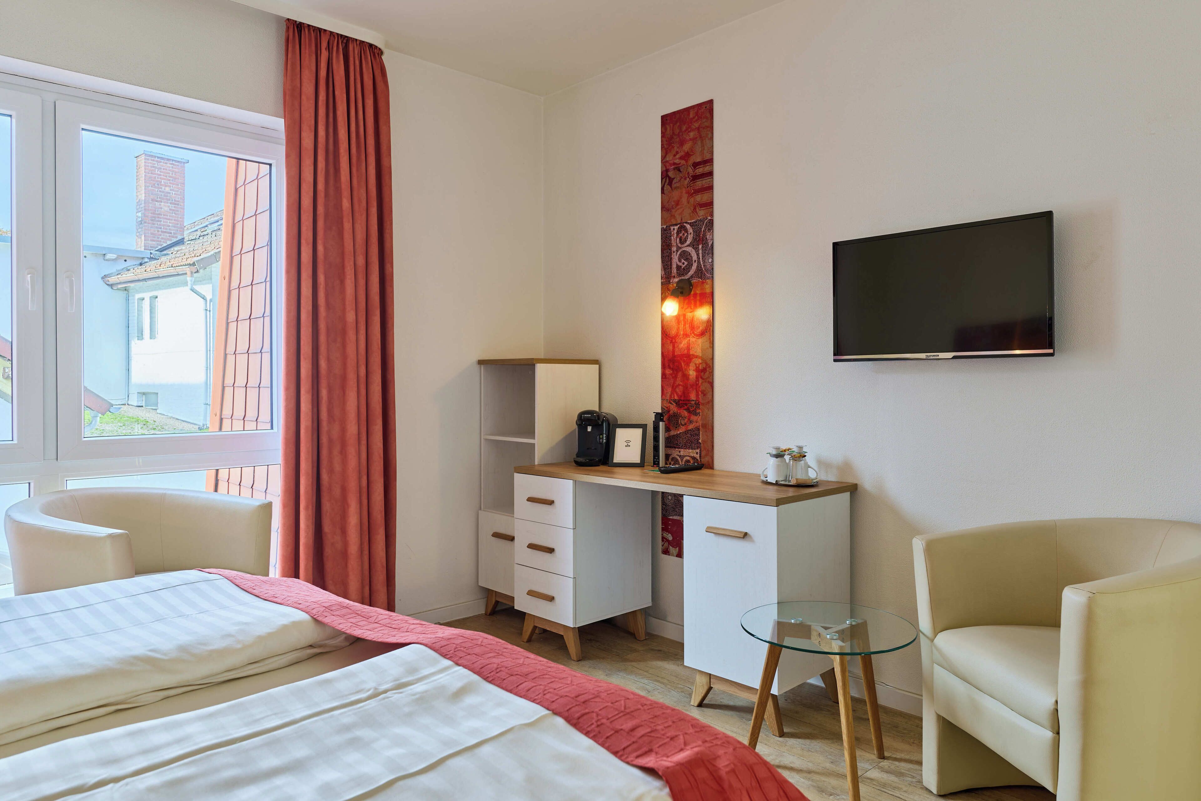 A hotel room with a bed, a desk with drawers, a chair, a TV mounted on the wall, and a window with curtains.