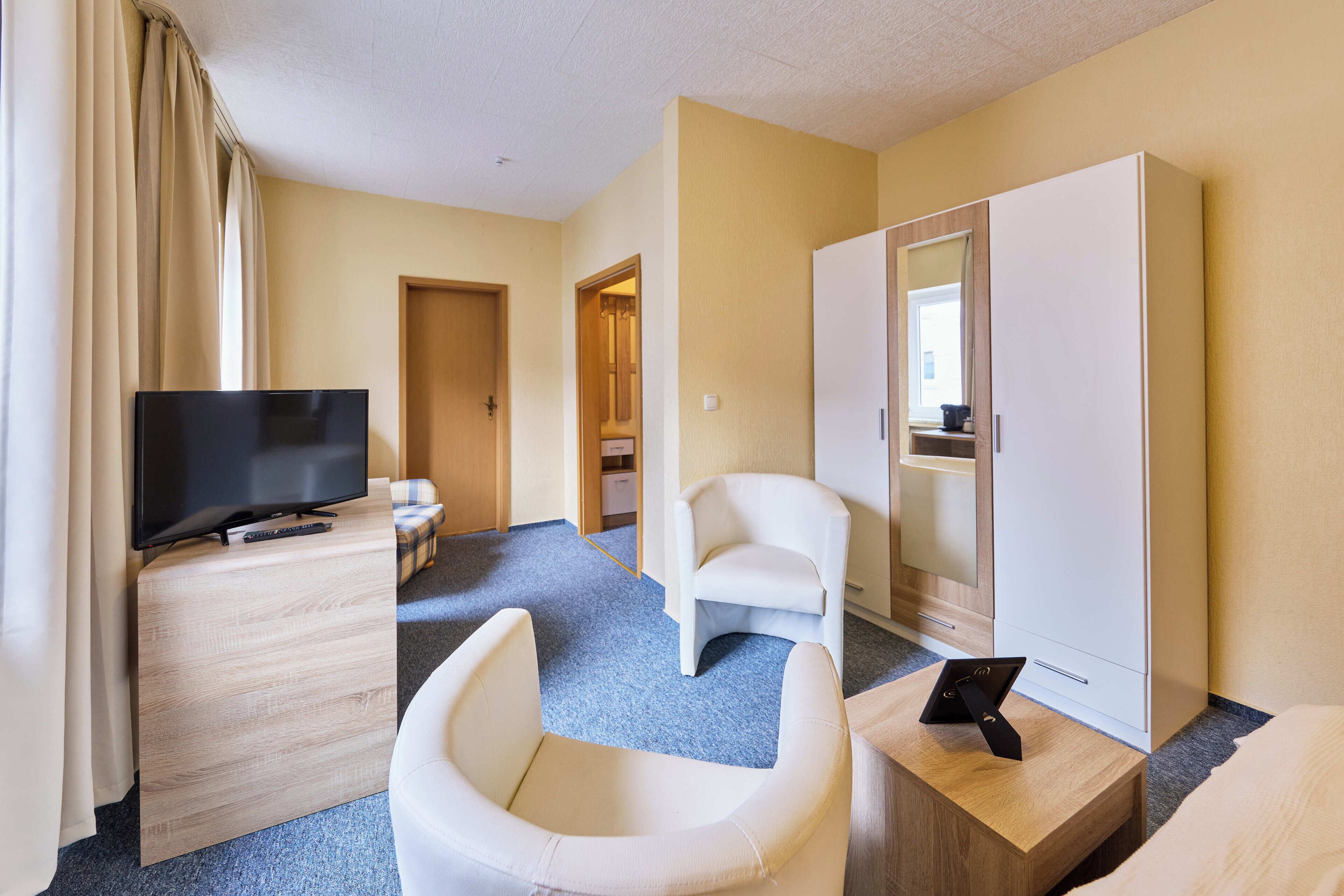 A hotel room with a flat-screen TV on a wooden side table, a white armchair, a sofa, and a wardrobe.