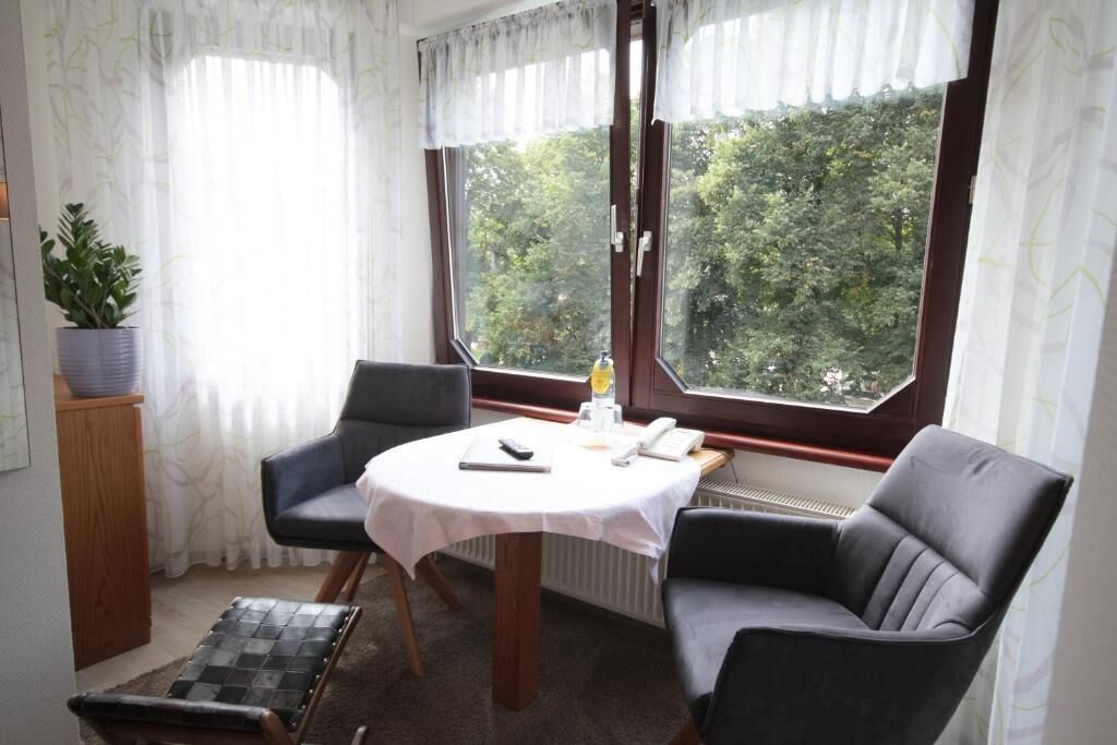 A room with a round table, two chairs, a radiator, a window with curtains, and a potted plant.